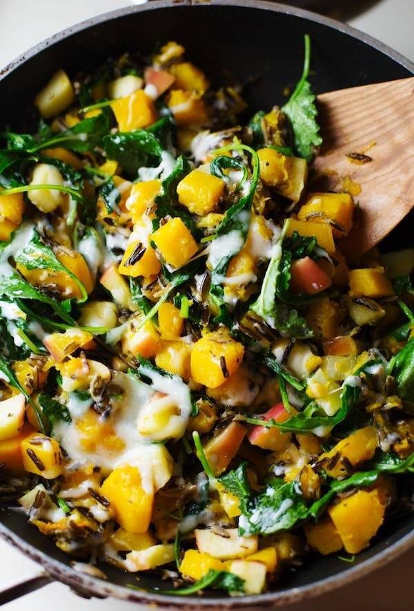 Apples, squash, sauteed onions, wild rice, and baby kale in a skillet with a wooden spoon.